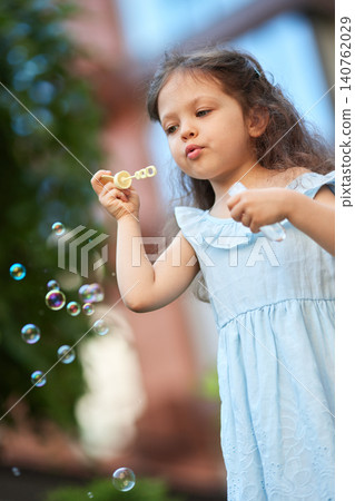 Girl in light blue dress blowing soap bubbles outdoors, symbolizing innocence, happiness, and carefree summer fun Girl in light blue dress blowing soap bubbles outdoors, symbolizing innocence, happiness, and carefree summer fun 140762029