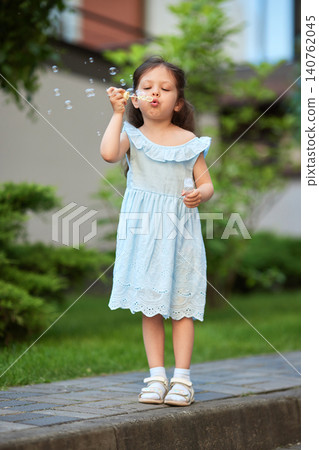 Young girl in a light blue sundress having fun blowing small soap bubbles while playing outside during summer Young girl in a light blue sundress having fun blowing small soap bubbles while playing outside during summer 140762045