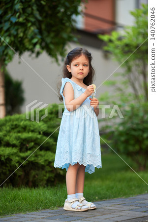 Little girl wearing a blue dress and white sandals standing on a paved path in a park holding a lollipop Little girl wearing a blue dress and white sandals standing on a paved path in a park holding a lollipop 140762046