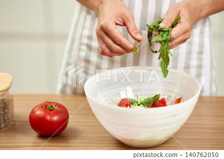 Person's hands adding green arugula leaves to a white bowl with chopped tomatoes and ingredients for a fresh meal Person's hands adding green arugula leaves to a white bowl with chopped tomatoes and ingredients for a fresh meal 140762050