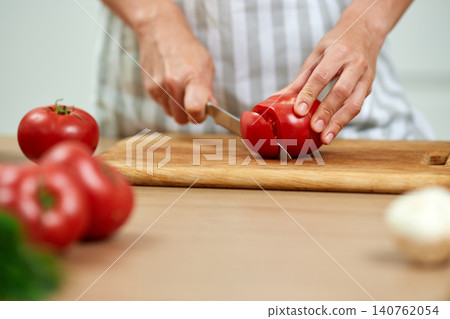 Hands slicing a fresh red tomato on a wooden cutting board, preparing healthy food in the kitchen Hands slicing a fresh red tomato on a wooden cutting board, preparing healthy food in the kitchen 140762054