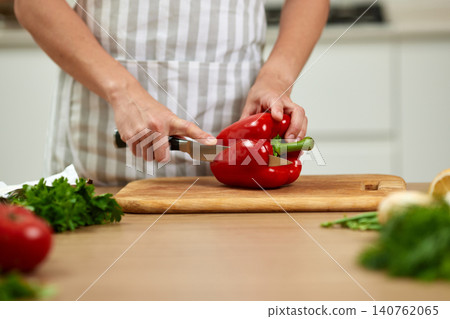 Person wearing an apron using a knife to slice a fresh red bell pepper on a rustic cutting board in a home kitchen Person wearing an apron using a knife to slice a fresh red bell pepper on a rustic cutting board in a home kitchen 140762065