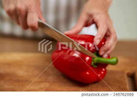 Hands preparing fresh red bell pepper with a chef's knife on a wooden cutting board, making healthy food Hands preparing fresh red bell pepper with a chef's knife on a wooden cutting board, making healthy food 140762066