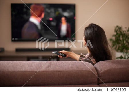 Woman sitting on a sofa in a modern living room, holding a remote control and watching tv, feeling comfortable 140762105