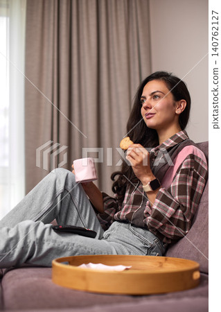 Young woman relaxing on sofa, enjoying a cookie and hot drink in her cozy living room at home. Snacking while watching tv Young woman relaxing on sofa, enjoying a cookie and hot drink in her cozy living room at home. Snacking while watching tv 140762127