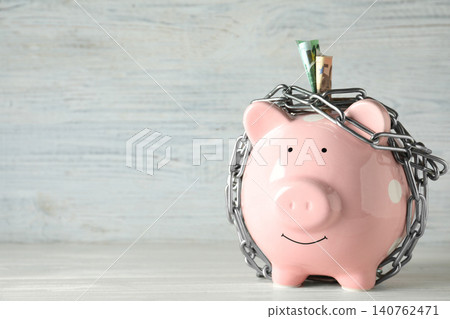 Piggy bank with steel chain and banknotes on white wooden table, space for text. Money safety concept Piggy bank with steel chain and banknotes on white wooden table, space for text. Money safety concept 140762471