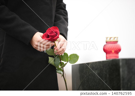 Woman holding red rose near black granite tombstone with candle outdoors, closeup. Funeral ceremony 140762595