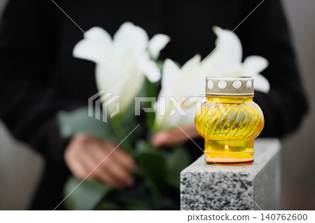 Woman holding white lilies near grey granite tombstone outdoors, focus on candle. Funeral ceremony Woman holding white lilies near grey granite tombstone outdoors, focus on candle. Funeral ceremony 140762600