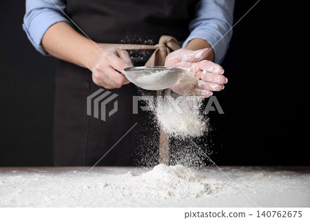 Woman sifting wheat flour at table against black background, closeup Woman sifting wheat flour at table against black background, closeup 140762675