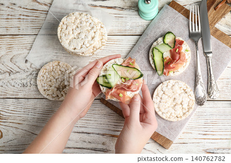 Woman holding puffed rice cake with prosciutto and cucumber at white wooden table, top view Woman holding puffed rice cake with prosciutto and cucumber at white wooden table, top view 140762782