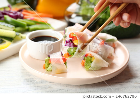 Woman eating delicious roll wrapped in rice paper at white wooden table, closeup Woman eating delicious roll wrapped in rice paper at white wooden table, closeup 140762790