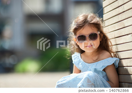 Little girl smiling, wearing stylish sunglasses and a blue dress, sitting outdoors on a warm sunny day 140762866