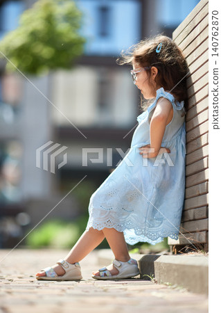 Young child in a light blue dress with glasses and an elaborate hem, leaning against a wooden wall on a summer day Young child in a light blue dress with glasses and an elaborate hem, leaning against a wooden wall on a summer day 140762870