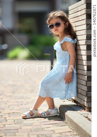 little child girl in a light blue sundress and sandals poses on a sunny urban sidewalk, playful and carefree on a warm summer day 140762871