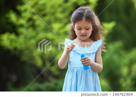 Young child in a blue dress playing outdoors with a bubble wand, enjoying childhood fun and summer activity Young child in a blue dress playing outdoors with a bubble wand, enjoying childhood fun and summer activity 140762904