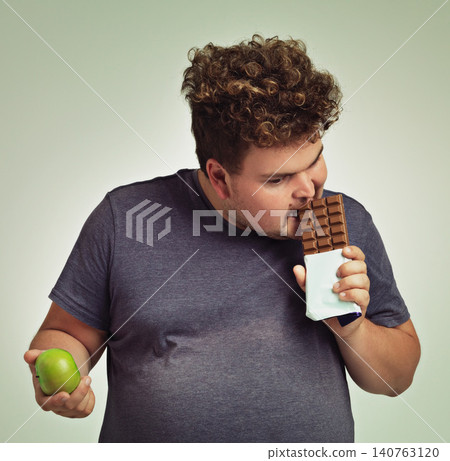 Man, apple and chocolate in studio with choice of junk food, sweet or diet for wellness. Plus size, male person eating and decision, fruit or candy for nutrition, lose weight and healthy lifestyle Man, apple and chocolate in studio with choice of junk food, sweet or diet for wellness. Plus size, male person eating and decision, fruit or candy for nutrition, lose weight and healthy lifestyle 140763120