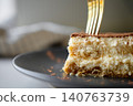 Fork presses into tiramisu slice on dark plate with cocoa topping and cream layers 140763739