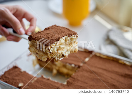 Hand lifts tiramisu slice with spatula from glass dish covered with cocoa powder Hand lifts tiramisu slice with spatula from glass dish covered with cocoa powder 140763740