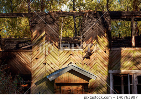 Charred remains of a rustic wooden house, its upper floor gutted by fire. Burnt timber contrasts with vibrant autumn trees seen through empty window frames. A poignant scene of decay Charred remains of a rustic wooden house, its upper floor gutted by fire. Burnt timber contrasts with vibrant autumn trees seen through empty window frames. A poignant scene of decay 140763997