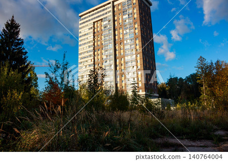 Tall modern apartment building peeking over wild autumn foliage under a vibrant blue sky, highlighting urban expansion near nature Tall modern apartment building peeking over wild autumn foliage under a vibrant blue sky, highlighting urban expansion near nature 140764004