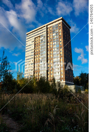 Modern high-rise residential building against a vibrant blue sky with fluffy clouds. Tall grasses frame the foreground, suggesting urban development meets wild nature Modern high-rise residential building against a vibrant blue sky with fluffy clouds. Tall grasses frame the foreground, suggesting urban development meets wild nature 140764005