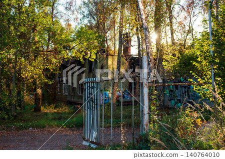An old, derelict wooden house, partially burnt, stands abandoned among vibrant autumn foliage. A rusty fence and street art frame this forgotten structure under warm sunlight An old, derelict wooden house, partially burnt, stands abandoned among vibrant autumn foliage. A rusty fence and street art frame this forgotten structure under warm sunlight 140764010