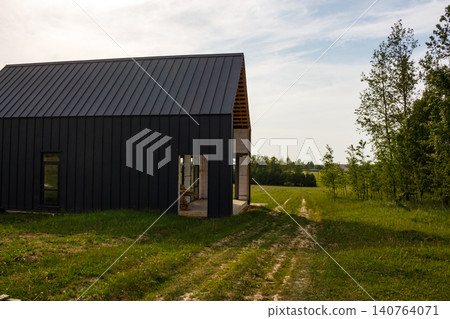 Modern barn-style house under construction, featuring dark siding and a sleek roof, nestled in a lush green field. A rustic path leads to distant trees under a bright sky 140764071