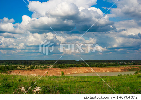 Rolling white and gray clouds dominate a vivid blue sky, overlooking a sandy quarry pond nestled in lush green fields and a distant forest 140764072