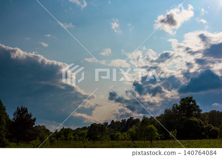 Dramatic cumulus clouds fill a vast summer sky, casting shadows over a serene green field and distant forest edge, hinting at an impending change in weather or late afternoon light Dramatic cumulus clouds fill a vast summer sky, casting shadows over a serene green field and distant forest edge, hinting at an impending change in weather or late afternoon light 140764084
