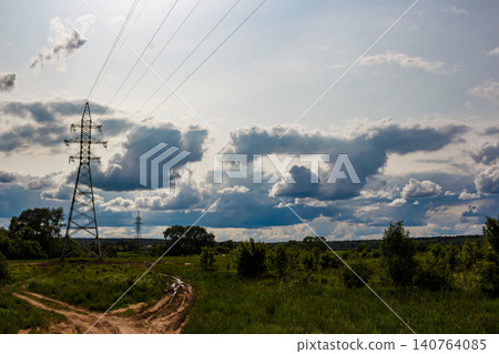 Sturdy electricity pylon dominates a vibrant green summer field under a dramatic, cloudy sky, with a dirt track leading into the distance Sturdy electricity pylon dominates a vibrant green summer field under a dramatic, cloudy sky, with a dirt track leading into the distance 140764085