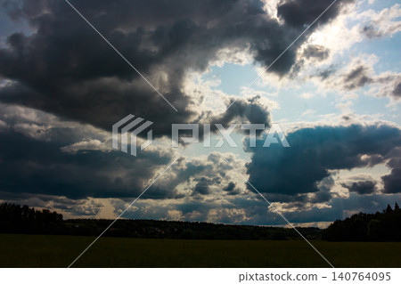 Intense, dramatic storm clouds dominate the sky, pierced by rays of light through gaps of blue. A moody sky over a tranquil green landscape, hinting at impending weather Intense, dramatic storm clouds dominate the sky, pierced by rays of light through gaps of blue. A moody sky over a tranquil green landscape, hinting at impending weather 140764095