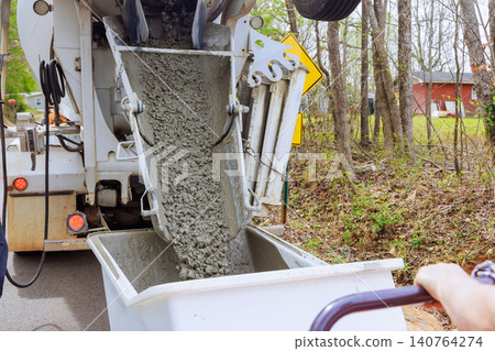 Worker guides concrete from truck into wheelbarrow dumper on construction site. Worker guides concrete from truck into wheelbarrow dumper on construction site. 140764274