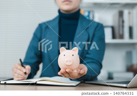 Piggy Bank and Financial Planning Concept. Businesswoman holding a pink piggy bank with a notebook and laptop on the desk, symbolizing financial planning, savings, investment, and money management. 140764331