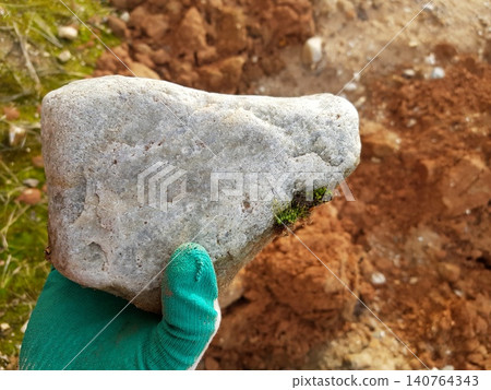 Close-up of a rough greenish quartzite boulder held by a gloved hand outdoors with raw earth in the background highlighting a unique geological find in a natural setting Close-up of a rough greenish quartzite boulder held by a gloved hand outdoors with raw earth in the background highlighting a unique geological find in a natural setting 140764343