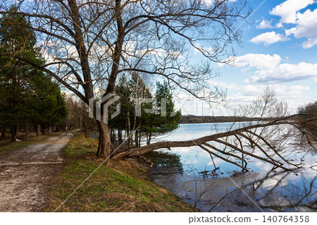 Spring flood overflowing a quiet country riverside, featuring a dramatic fallen tree stretching over tranquil waters under a bright blue sky with soft white clouds, early April seasonal landscape Spring flood overflowing a quiet country riverside, featuring a dramatic fallen tree stretching over tranquil waters under a bright blue sky with soft white clouds, early April seasonal landscape 140764358