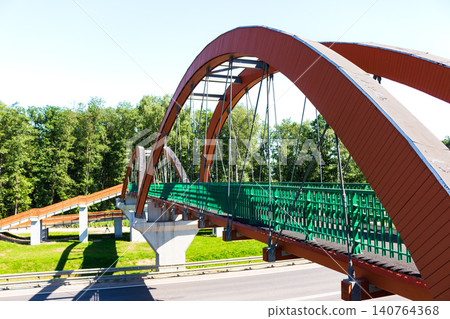 Modern red arched pedestrian bridge spanning over a highway, featuring a metal frame and vibrant green railings, set against a backdrop of lush green summer forest trees Modern red arched pedestrian bridge spanning over a highway, featuring a metal frame and vibrant green railings, set against a backdrop of lush green summer forest trees 140764368