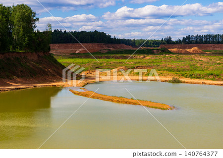 Scenic view of a tranquil turquoise pond nestled within a vast open clay pit quarry surrounded by wild green forest hills under a soft cloudy summer sky 140764377