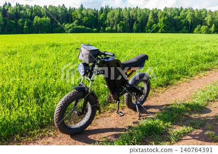 Black electric bike parked on a dirt path near a lush green field during a sunny summer day, modern eco-friendly transport for adventure and off-road riding in nature Black electric bike parked on a dirt path near a lush green field during a sunny summer day, modern eco-friendly transport for adventure and off-road riding in nature 140764391