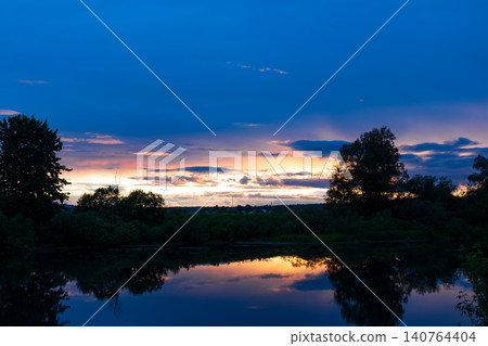 Vibrant sunset paints the sky with golden hues above a calm river reflecting dense silhouettes of trees in the fading evening light, capturing a peaceful and serene nature moment Vibrant sunset paints the sky with golden hues above a calm river reflecting dense silhouettes of trees in the fading evening light, capturing a peaceful and serene nature moment 140764404