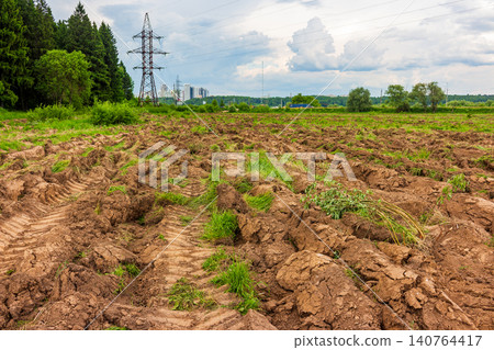 Freshly plowed overgrown field with deep tractor tire tracks, showing raw brown earth and patches of green grass under a cloudy sky in a rural agricultural landscape Freshly plowed overgrown field with deep tractor tire tracks, showing raw brown earth and patches of green grass under a cloudy sky in a rural agricultural landscape 140764417