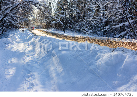 Snowy forest road after a blizzard with high white drifts and a lone pedestrian walking along the path under bright sunny winter sky near the frozen woodland canopy Snowy forest road after a blizzard with high white drifts and a lone pedestrian walking along the path under bright sunny winter sky near the frozen woodland canopy 140764723