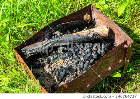 Rusty disposable metal grill left in green grass with burnt logs and charcoal ashes, an example of irresponsible camping and environmental pollution in a summer meadow setting 140764737