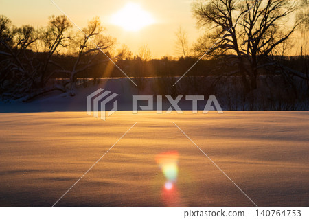 Golden sunset light casts a warm glow over a pristine snow-covered field with lens flare effects, framed by dark silhouettes of winter trees against a serene evening sky Golden sunset light casts a warm glow over a pristine snow-covered field with lens flare effects, framed by dark silhouettes of winter trees against a serene evening sky 140764753