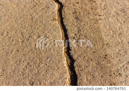 Deep longitudinal fracture splitting old weathered concrete pavement on a rural road, textured dusty surface with earthy tones, top-down perspective showcasing structural damage and decay Deep longitudinal fracture splitting old weathered concrete pavement on a rural road, textured dusty surface with earthy tones, top-down perspective showcasing structural damage and decay 140764761