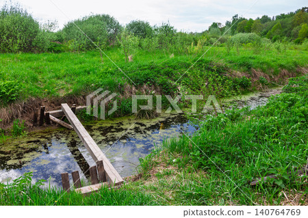 Rustic wooden plank bridge spanning a stagnant overgrown creek amidst a lush wild meadow with green bushes and young trees under a cloudy spring sky in the countryside Rustic wooden plank bridge spanning a stagnant overgrown creek amidst a lush wild meadow with green bushes and young trees under a cloudy spring sky in the countryside 140764769