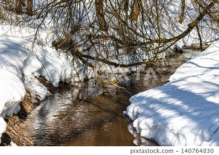 Bubbling spring stream with melting snowbanks along its winding path. Bare branches frame this vibrant scene of nature's thaw and reawakening 140764830