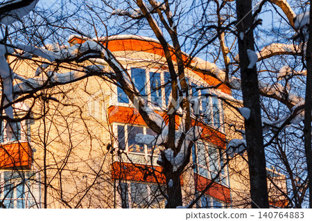 Golden-lit apartment building peeks through a frosty winter forest canopy, bare branches heavy with fresh snow under a crisp blue sky Golden-lit apartment building peeks through a frosty winter forest canopy, bare branches heavy with fresh snow under a crisp blue sky 140764883