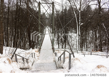 Pedestrian suspension bridge blanketed in fresh snow, spanning a serene winter forest. Gentle flakes fall, creating a quiet, picturesque scene Pedestrian suspension bridge blanketed in fresh snow, spanning a serene winter forest. Gentle flakes fall, creating a quiet, picturesque scene 140764888