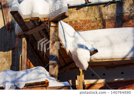 Rustic wooden scaffolding heavily laden with thick snow drifts and hanging icicles against a sunny brick wall backdrop 140764890