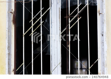Dilapidated window frame with security bars and shattered glass panes, showing decay and neglect behind dark interior 140764948